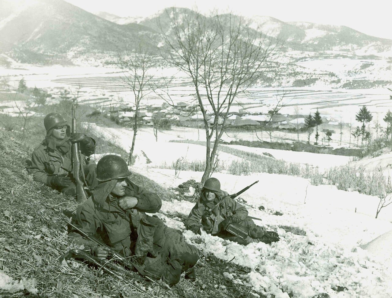 Three men bundled in winter gear sit on the side of a snow-covered hill.
