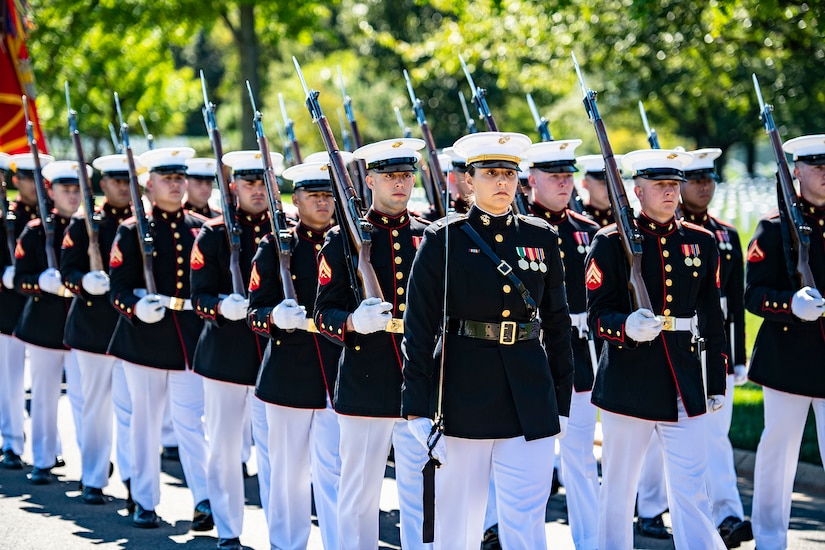 Marines in dress uniform stand in three lines while carrying rifles over their shoulders.