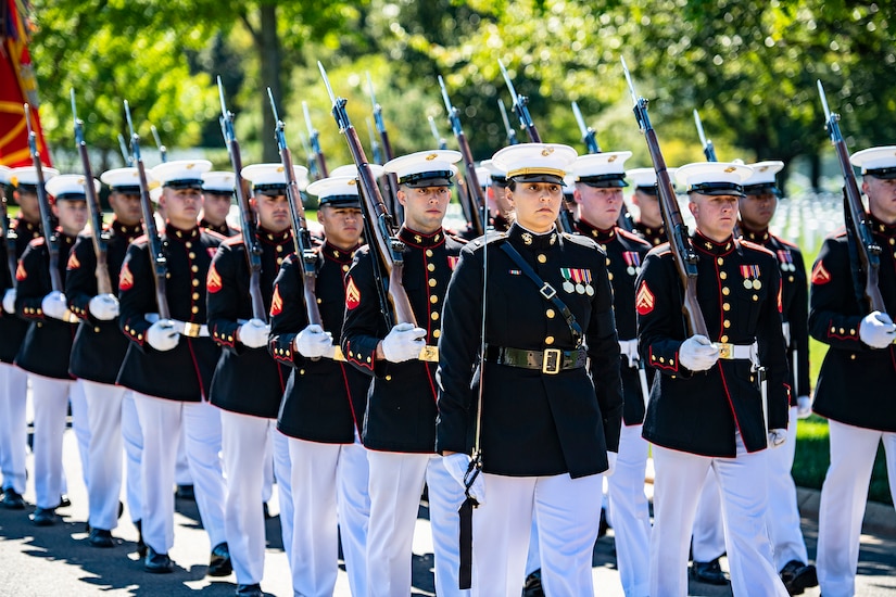 A woman in dress uniform looks stoically ahead while carrying a sword over her shoulder.