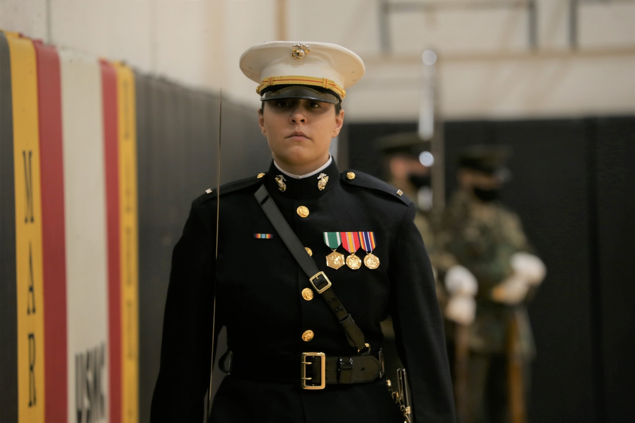 A woman in dress uniform looks stoically ahead while carrying a sword over her shoulder.