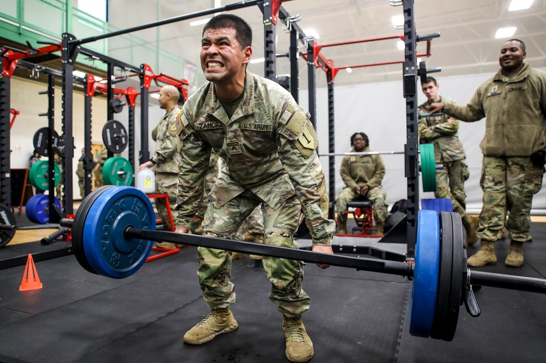 A soldier in a weight room lifts a barbell as others watch.