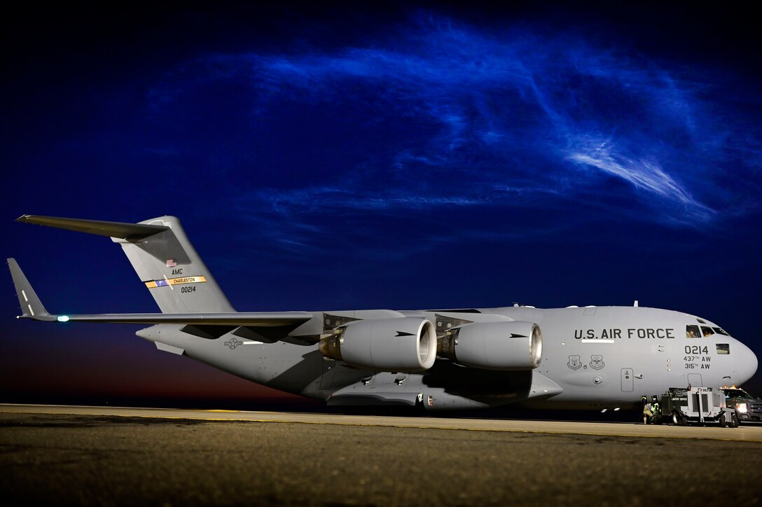 Steaks of light illuminate a dark blue sky above an aircraft parked on a flight line.