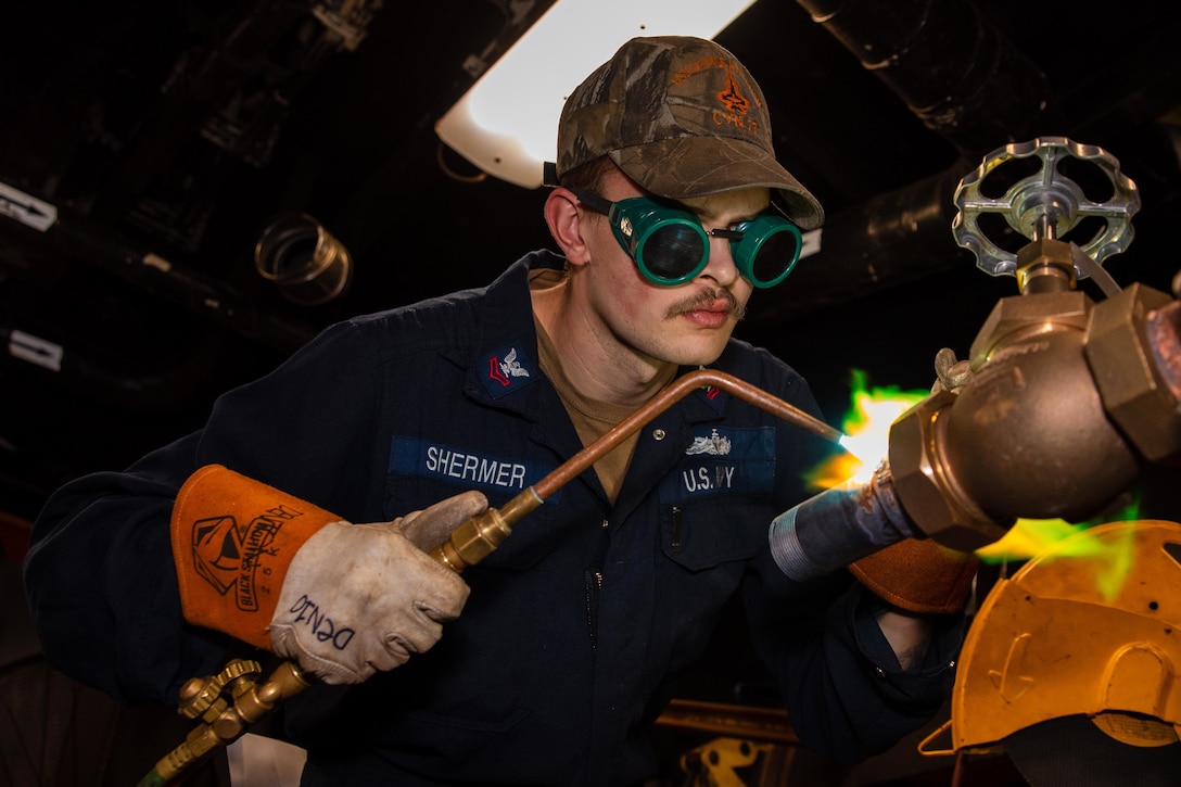 A sailor fires a welding torch against a seawater supply line on a ship.