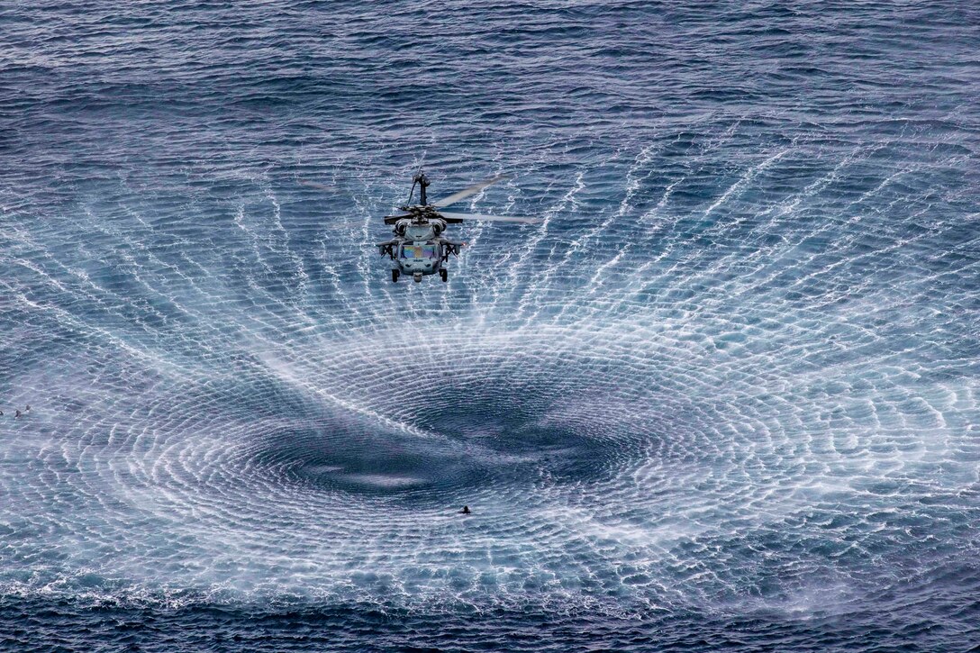 A helicopter creates a circular wake while hovering above a sailor in the water.