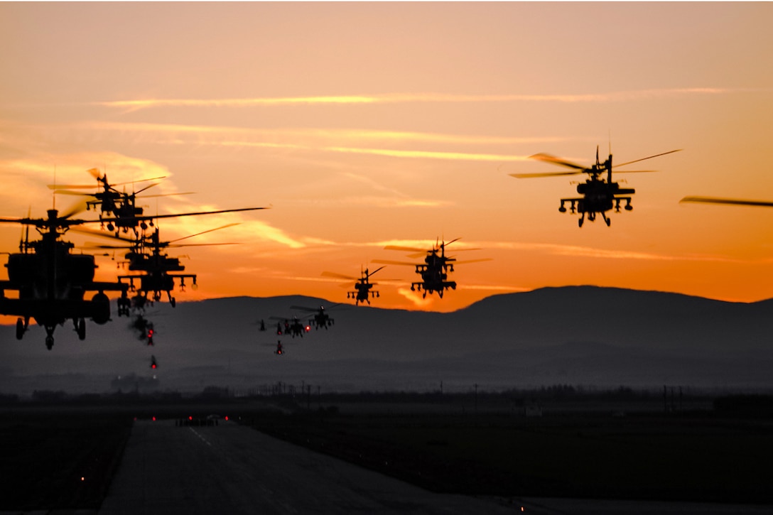 Helicopters fly above a runway against an orange sky.