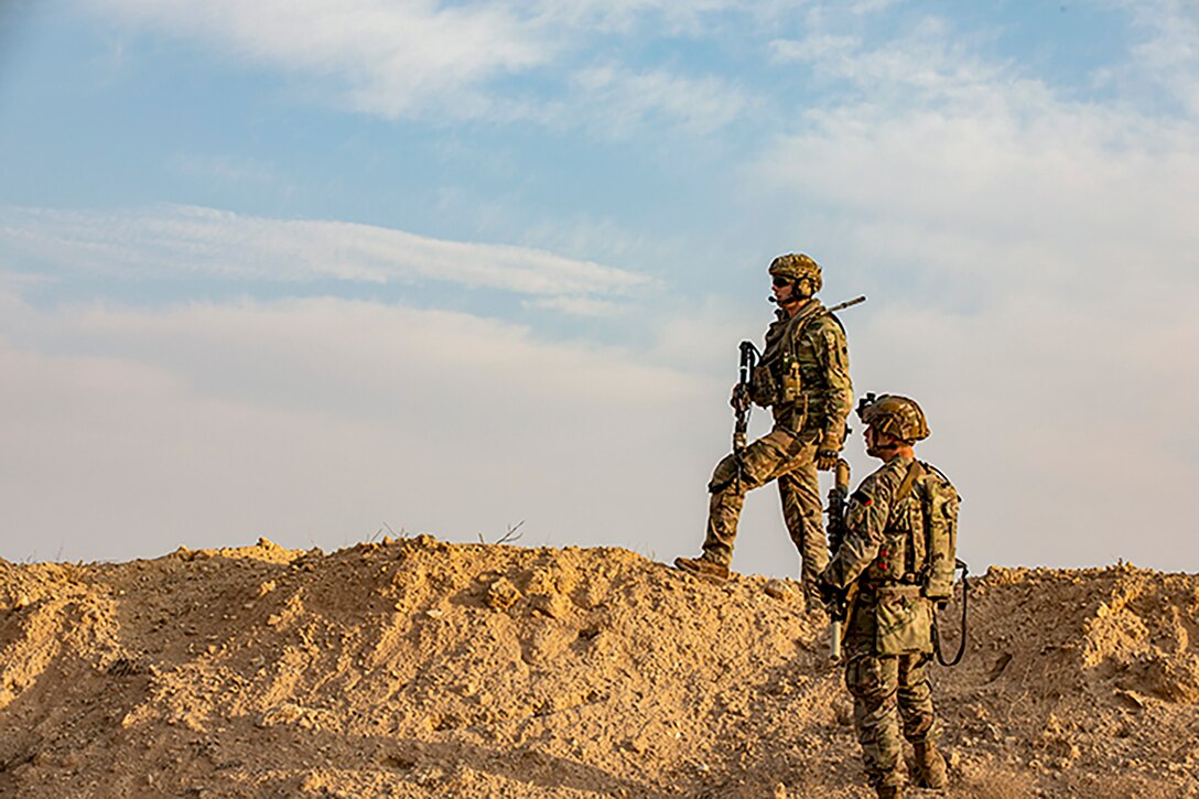 A soldier stands on a dirt mound by another soldier, both looking into the distance.