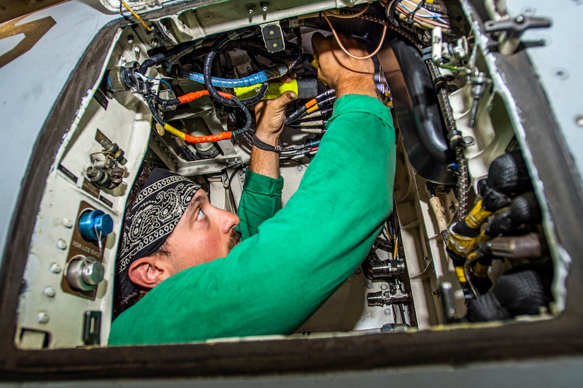 A sailor handles wires inside an aircraft frame.