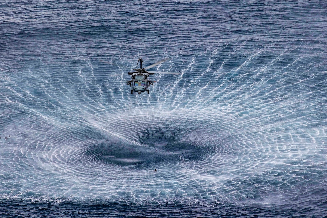 A helicopter creates a circular wake while hovering above a sailor in the water.