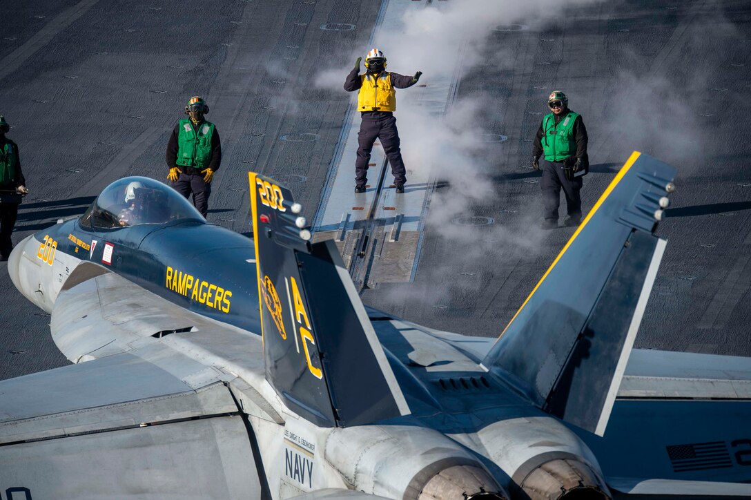 Sailors stand on a ship's flight deck, looking toward a military aircraft.