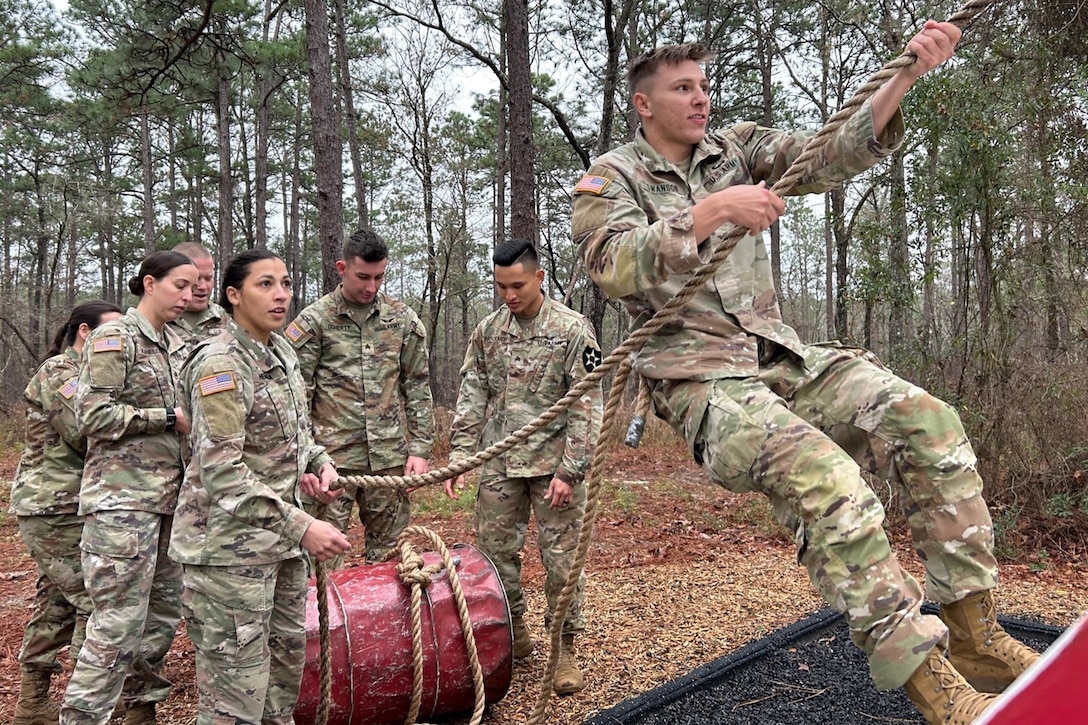 Soldiers watch as a fellow soldier climbs a rope.