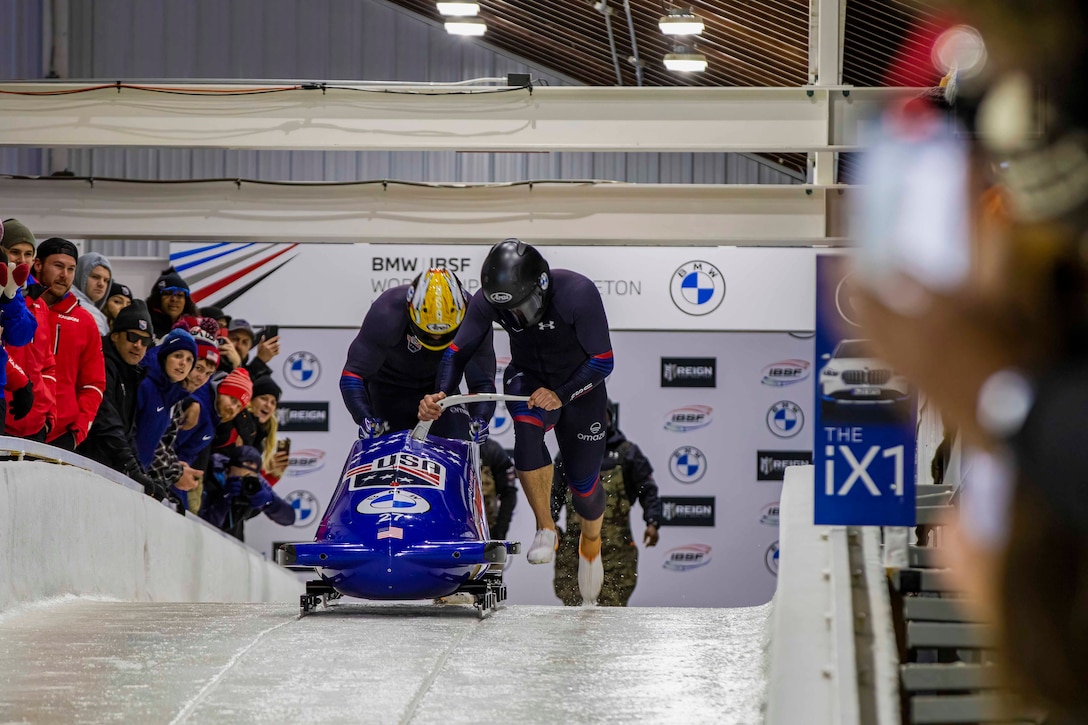 Two soldiers run with a bobsled on ice in front of a crowd.