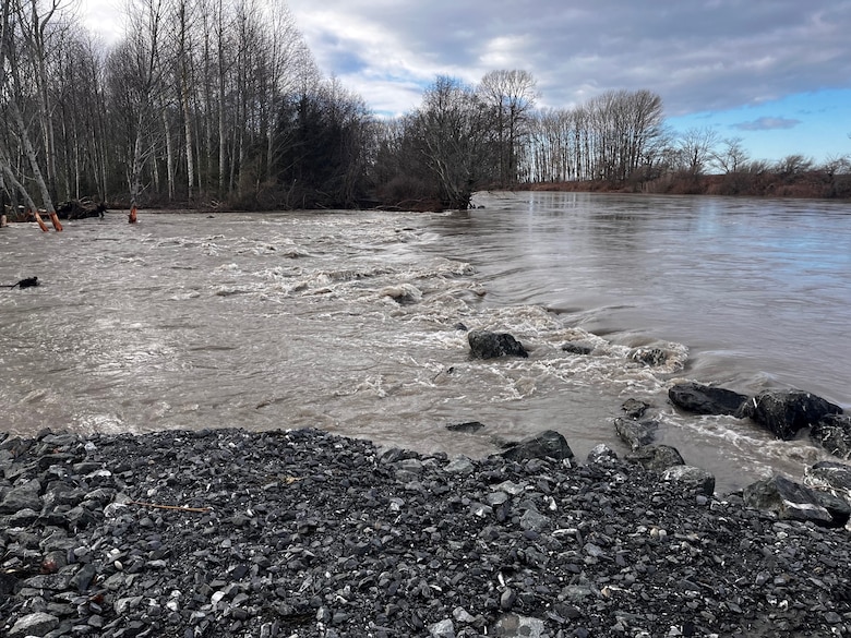 Outdoor scene with partly cloudy sky and trees in the background. Foreground shows water from the Nooksack river flowing over and through a levee breach