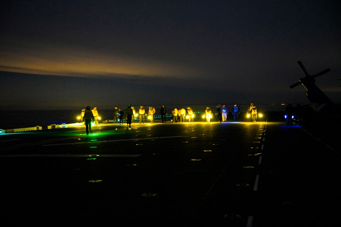 A line of sailors walk on the flight deck of a ship at night.