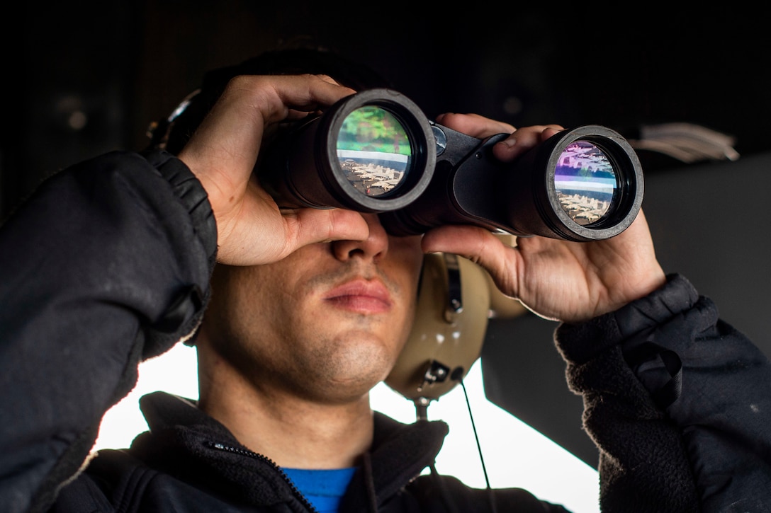 A sailor holds binoculars up to his eyes.