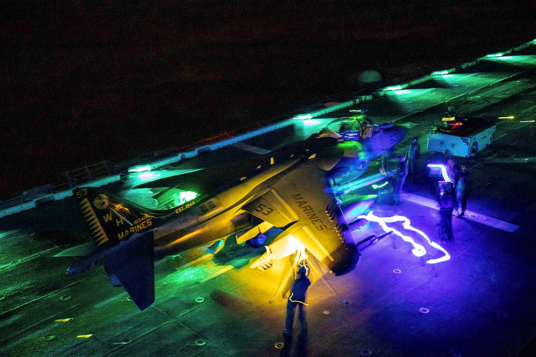 Sailors work on an aircraft aboard a ship at sea illuminated by colorful lights.