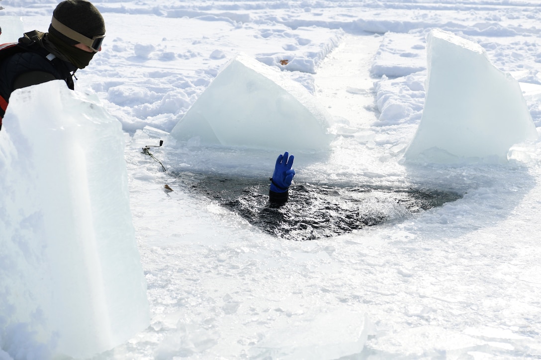 A service member holds their hand up while submerged in icy water surrounded by snow as a fellow service member watches.