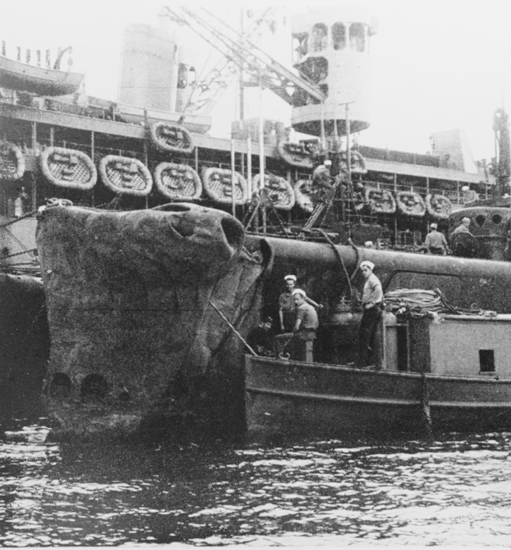 Three sailors sit in a small boat alongside a damaged submarine, which is next to a larger ship.