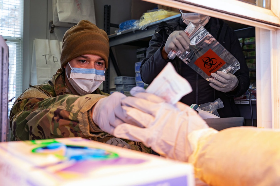 An airman wearing a face mask and gloves is handed a COVID-19 test specimen by someone wearing gloves and a gown.
