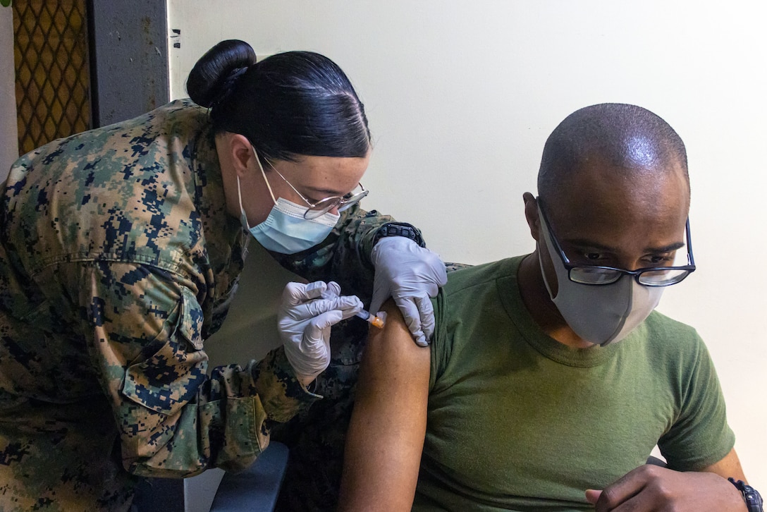 A Marine wearing a face mask is given a COVID-19 booster shot while seated by another Marine wearing a face mask and gloves who leans over to administer the vaccine.