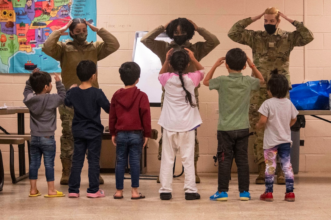 Three  airmen wearing face masks touch their heads as they sing a song with Afghan children.