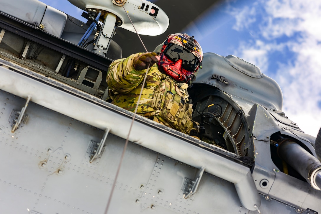 An airman wearing helmet looks out of an airborne helicopter while holding onto a rope.