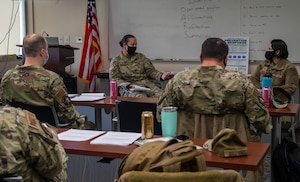 People sit in a classroom watch two women speaking each other at the front of the room