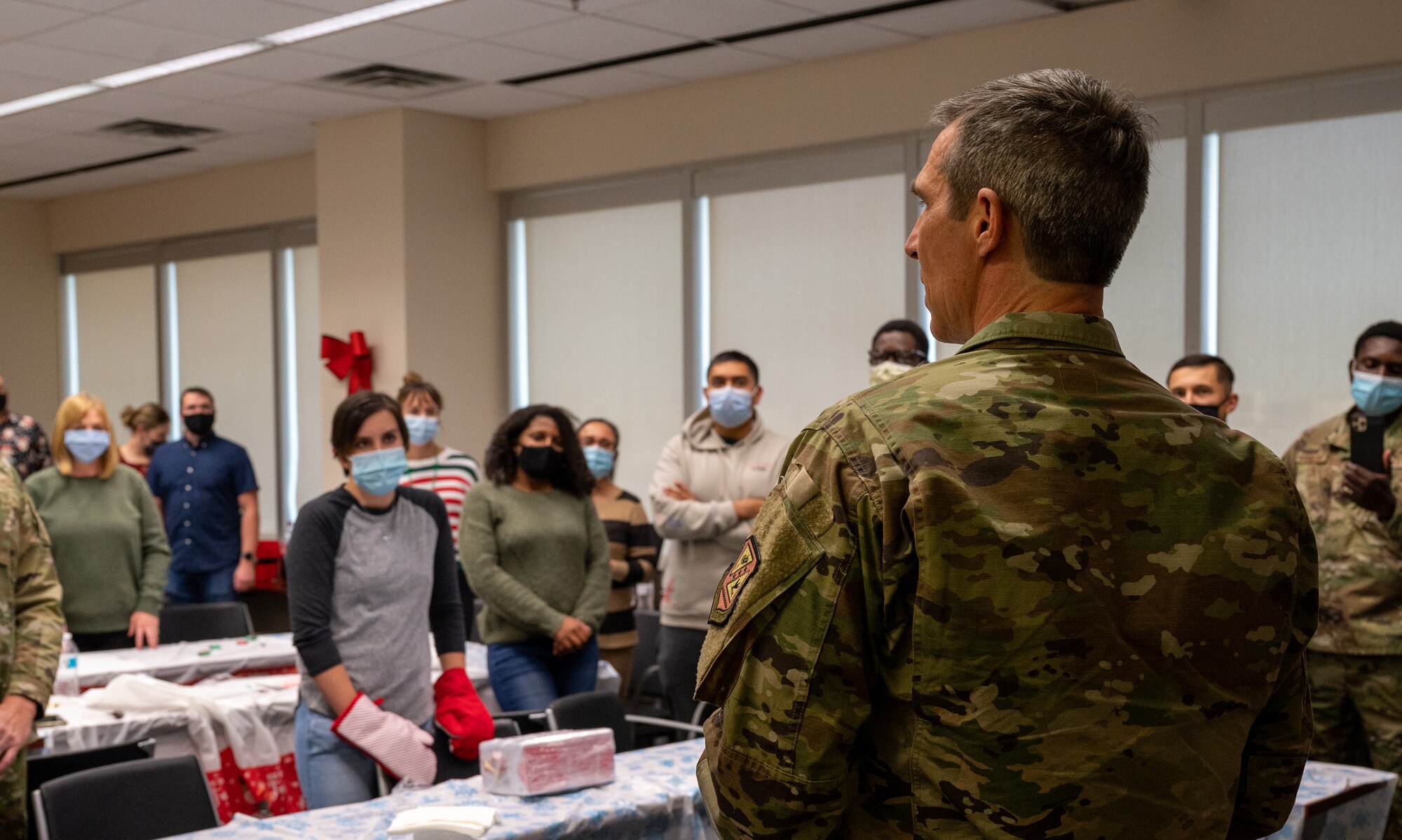 A man stands in a windowed room speaking to a group of people wearing facemasks.