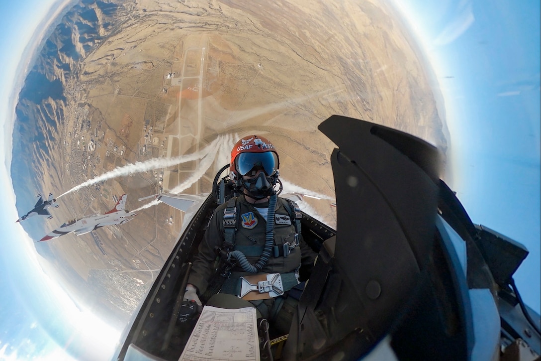 A pilot flying a military jet looks into the camera as other jets fly behind him.