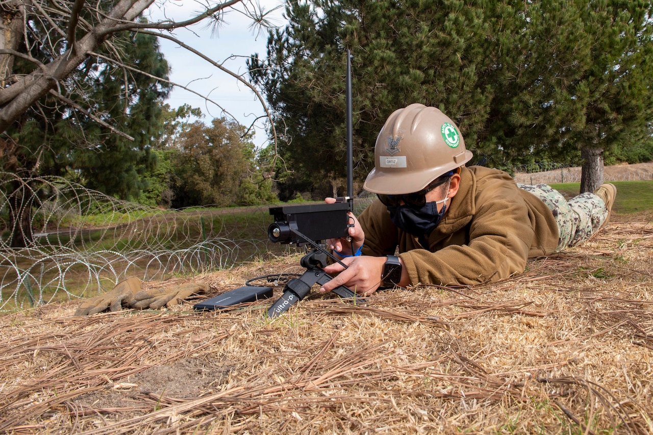 A service member lies on her stomach in the grass and points an electronic device ahead.