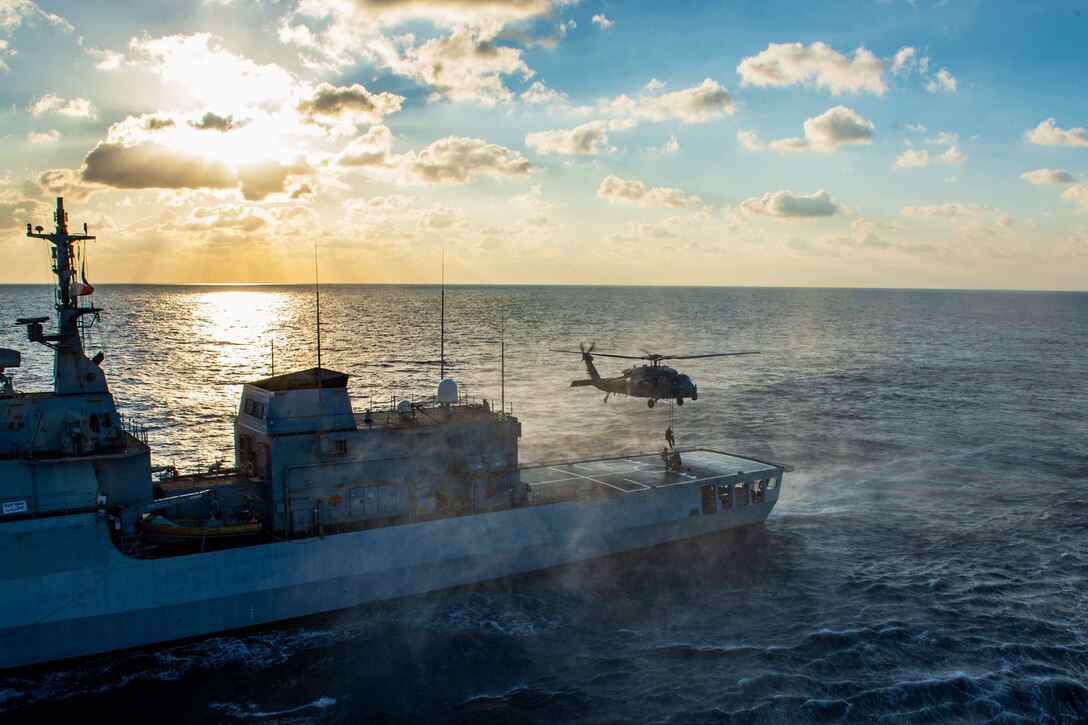 Sailors fast rope onto an Italian ship while transiting a body of water under a sunlit sky.