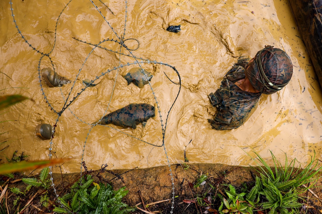 A Marine is seen from above laying in muddy water beneath barbed wire.