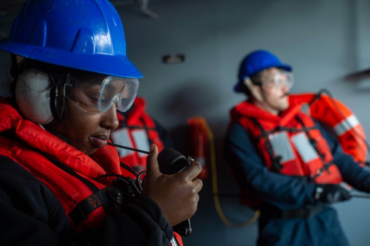 Sailors wearing orange flotation devices use a phone to communicate on a ship.