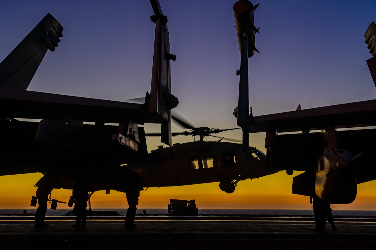 A helicopter lifts off from behind other aircraft on a flight deck.