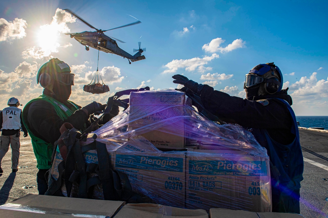 Two sailors remove wrapping from a pallet of supplies on the deck of a ship while a helicopter drops more in the background.