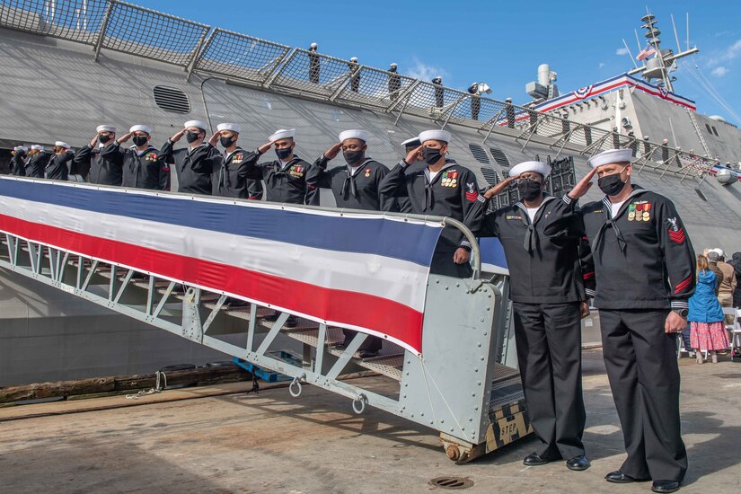 Sailors salute while standing in a row on a metal ramp leading from dock to ship.