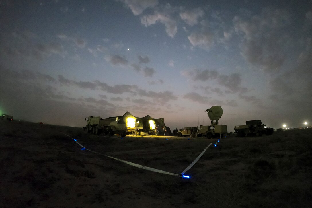 Expandable van shelters are parked in a field illuminated by blue lights.