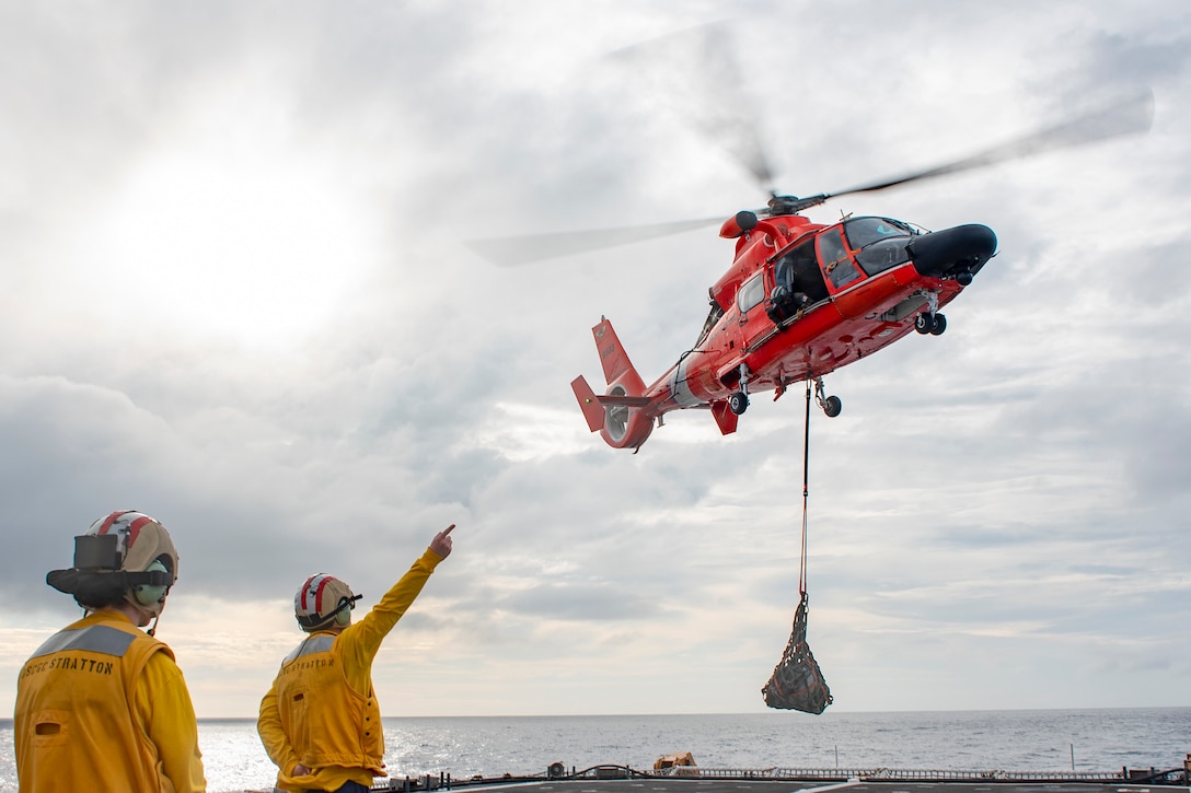 A Coast Guardsman raises his arm towards a hovering helicopter.