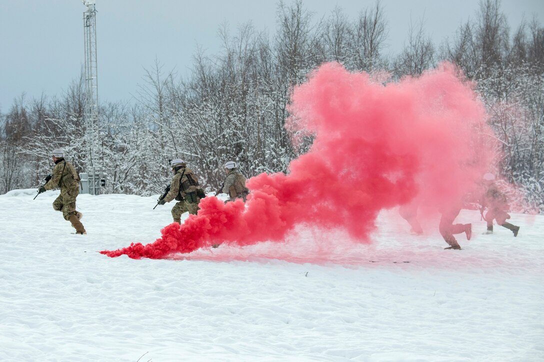 Paratroopers run across the snow as a pink smoke  rises from the ground.