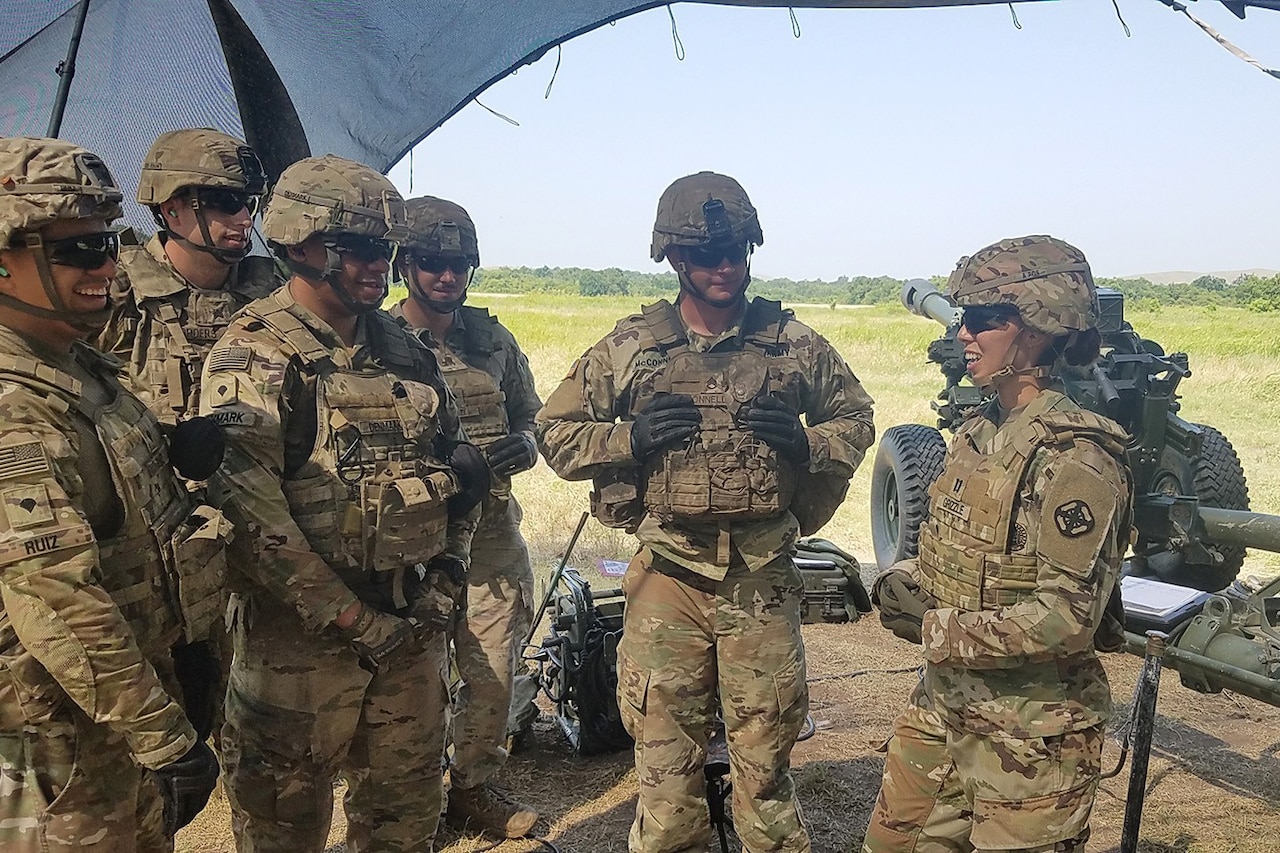 A soldier addresses other soldiers under mesh netting in a field.