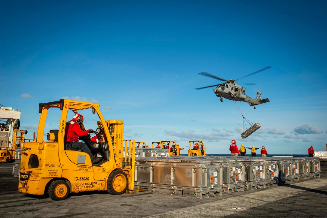 A group of sailors signal a hovering helicopter with dangling cargo while a yellow vehicle moves delivered containers.