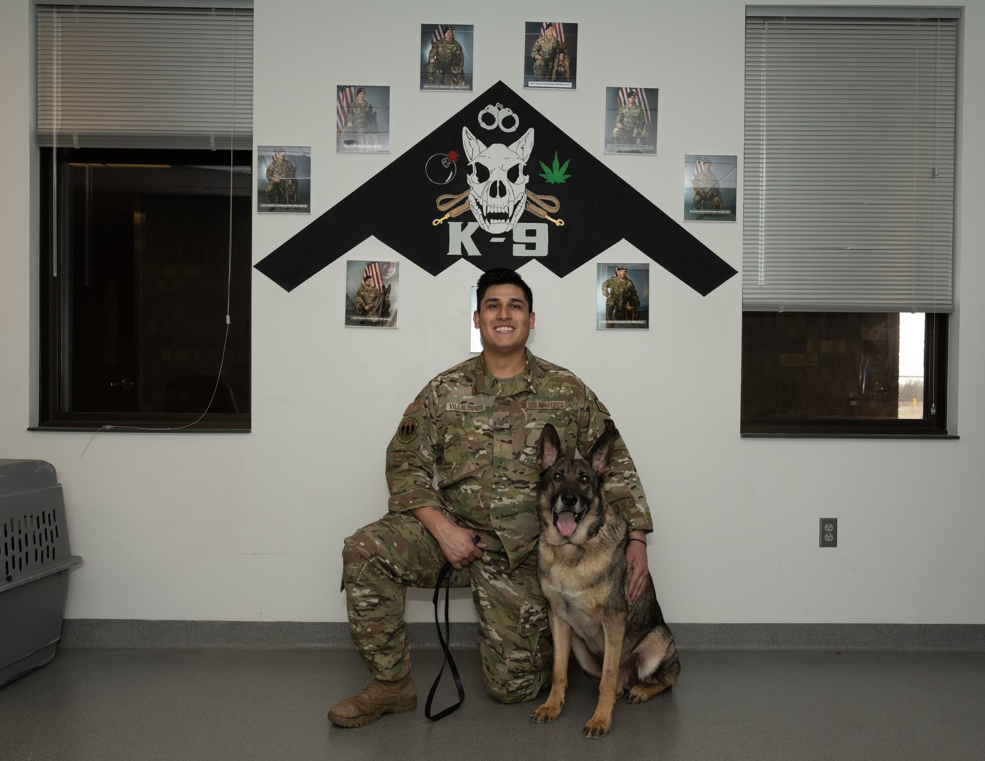 U.S. Air Force Staff Sgt. Alex Villalpando, 509 Security Forces military working dog handler, poses for a photo with MWD Stiffler in front of a wall showcasing military working dogs and their handlers at the MWD kennels on Whiteman Air Force Base Jan. 19, 2022. Military working dogs provide a valuable force multiplier to aid in security and readiness within our units. (U.S. Air Force photo by Airman 1st Class Bryson Britt)