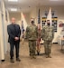 Man in suit and two Soldiers in uniform stand in recruiting office in front of Army flag and American flag.