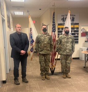 Man in suit and two Soldiers in uniform stand in recruiting office in front of Army flag and American flag.