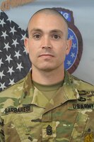 A man in an army uniform standing in front of 2 flags.