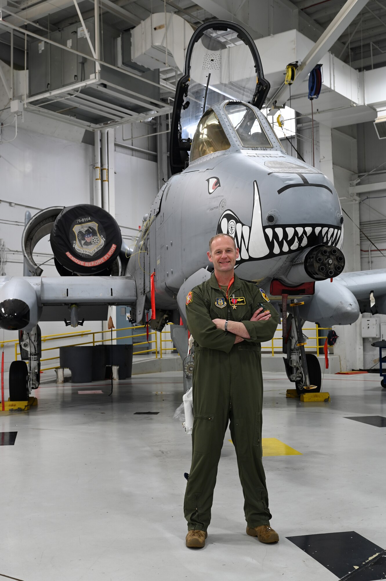 A man in an olive-green flight suit stands with arms folded in front of an A-10 Thunderbolt II in a hangar. A Bronze Star hangs from the man's chest.