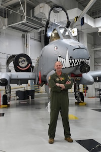 A man in an olive-green flight suit stands with arms folded in front of an A-10 Thunderbolt II in a hangar. A Bronze Star hangs from the man's chest.
