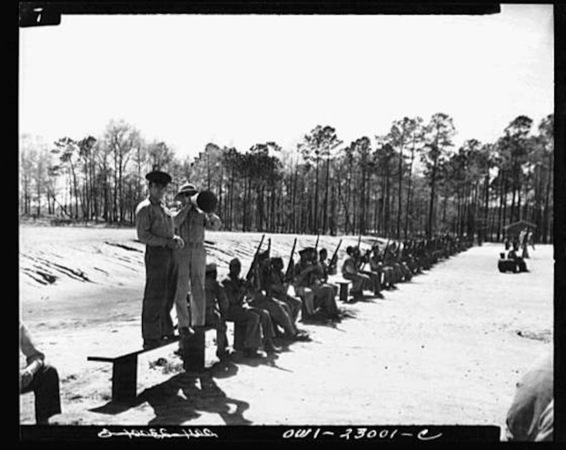 Marines sit in a row with their rifles as two men stand near them.