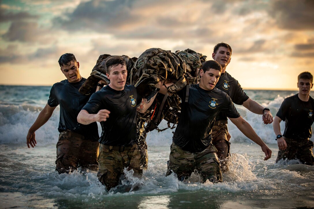 Soldiers carry a large bag while walking in a body of water under a gold and cloudy sky.