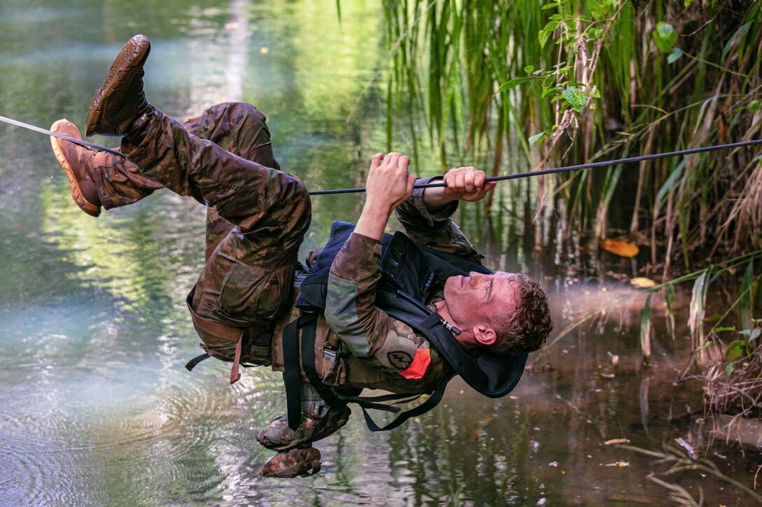 A soldier hoists himself across a rope bridge over water.