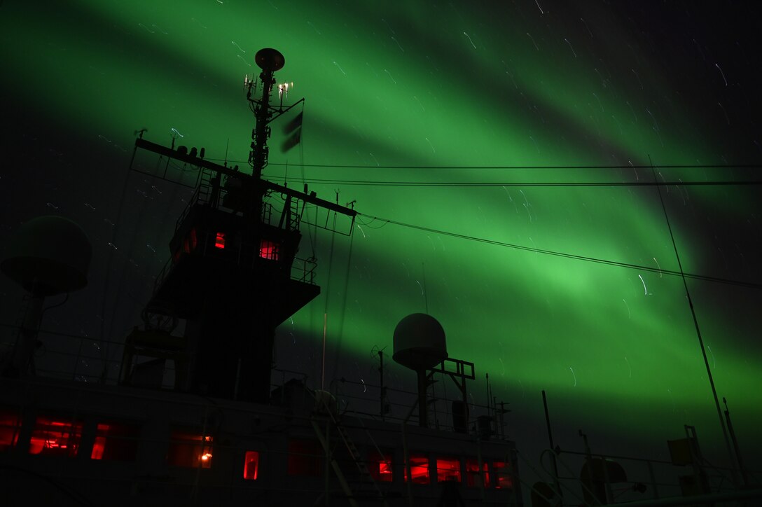 A green light in the sky backlights the cables of a ship in silhouette.